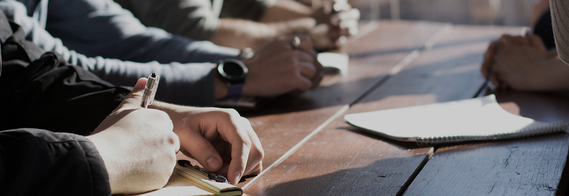 Personen unterhalten sich am Tisch, einer notiert etwas.